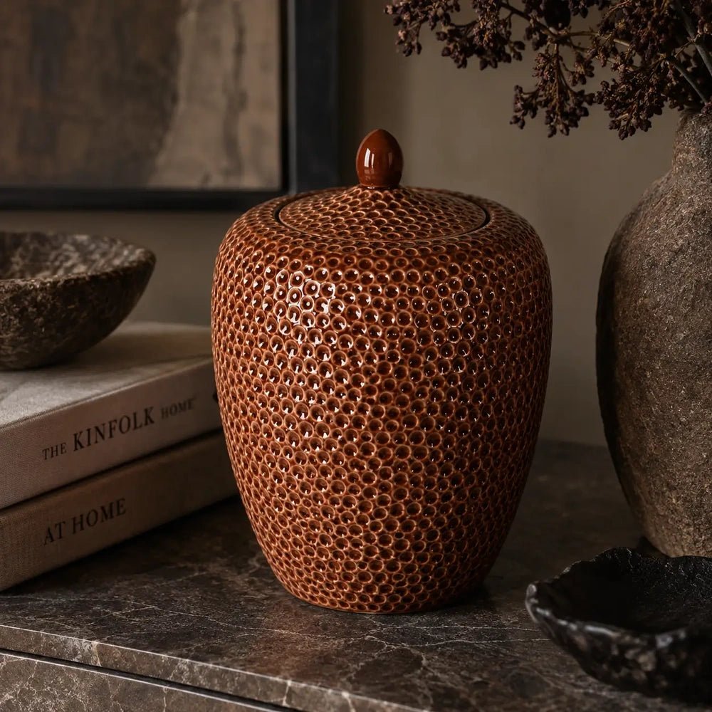 Nara Ceramic Jar in terracotta features a rounded lidded shape and a glossy dimpled surface, styled on a dark console beside books and sculptural accessories.