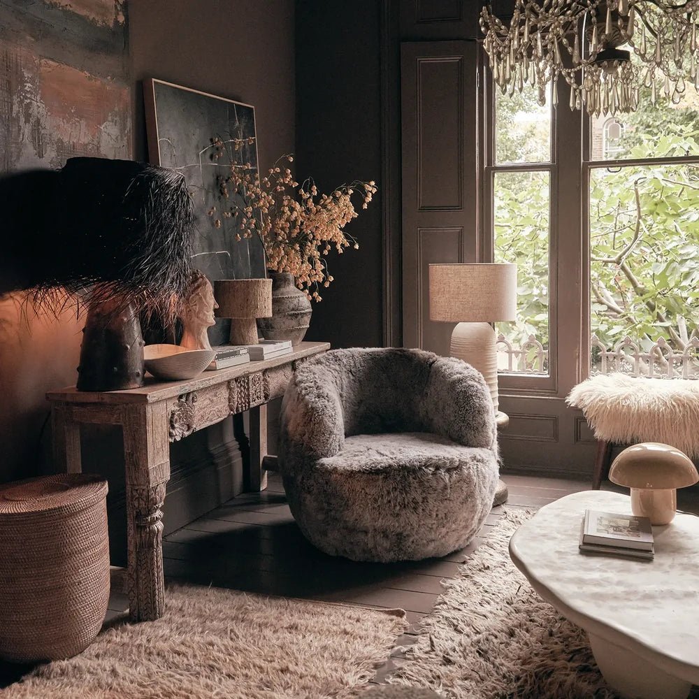 Norwood Sheepskin Swivel Chair features grey textured upholstery and a curved seat, positioned beside a wooden console beneath tall windows in a layered living room.
