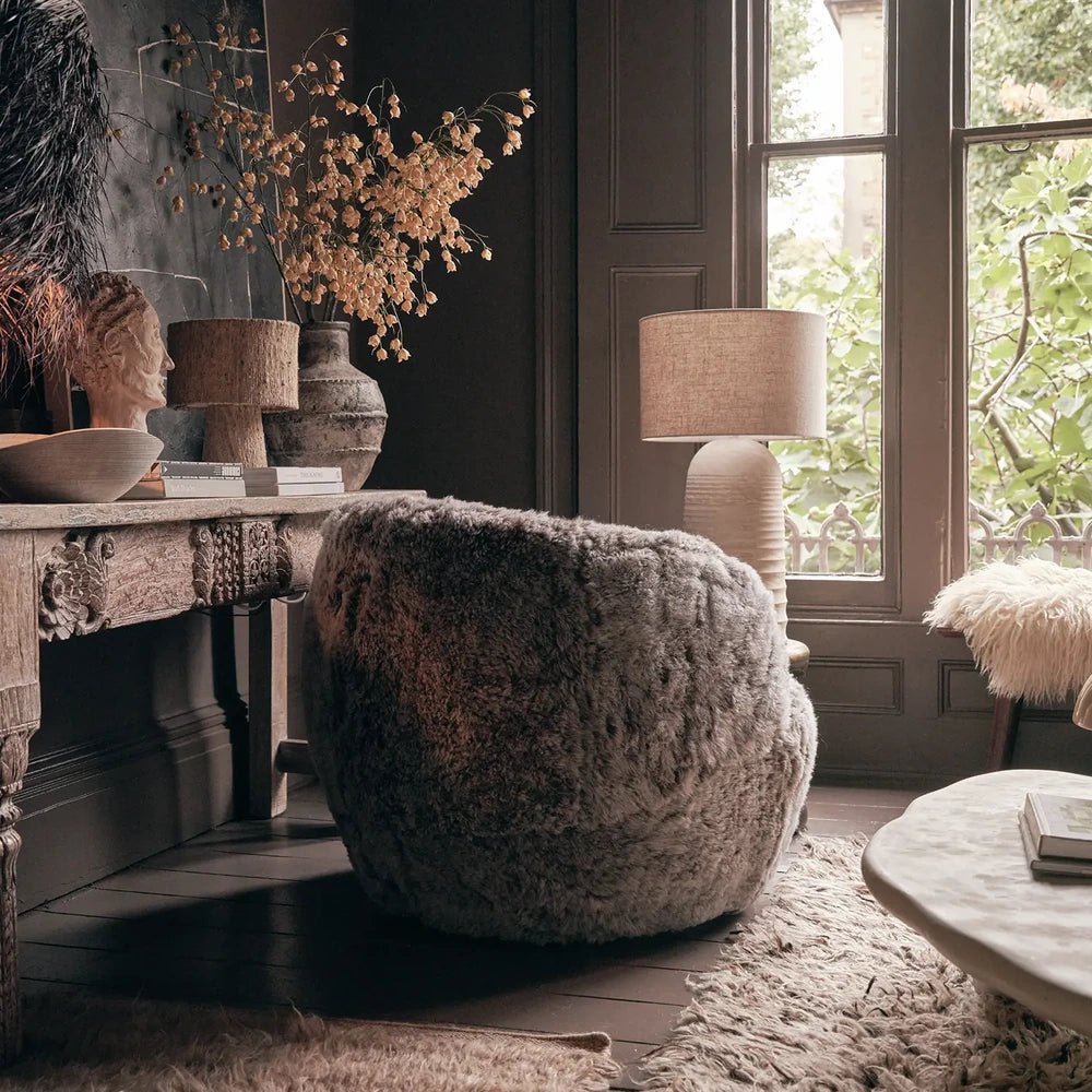 Norwood Sheepskin Swivel Chair reveals a rounded grey upholstered back, positioned beside a wooden console and tall window with a textured rug beneath.