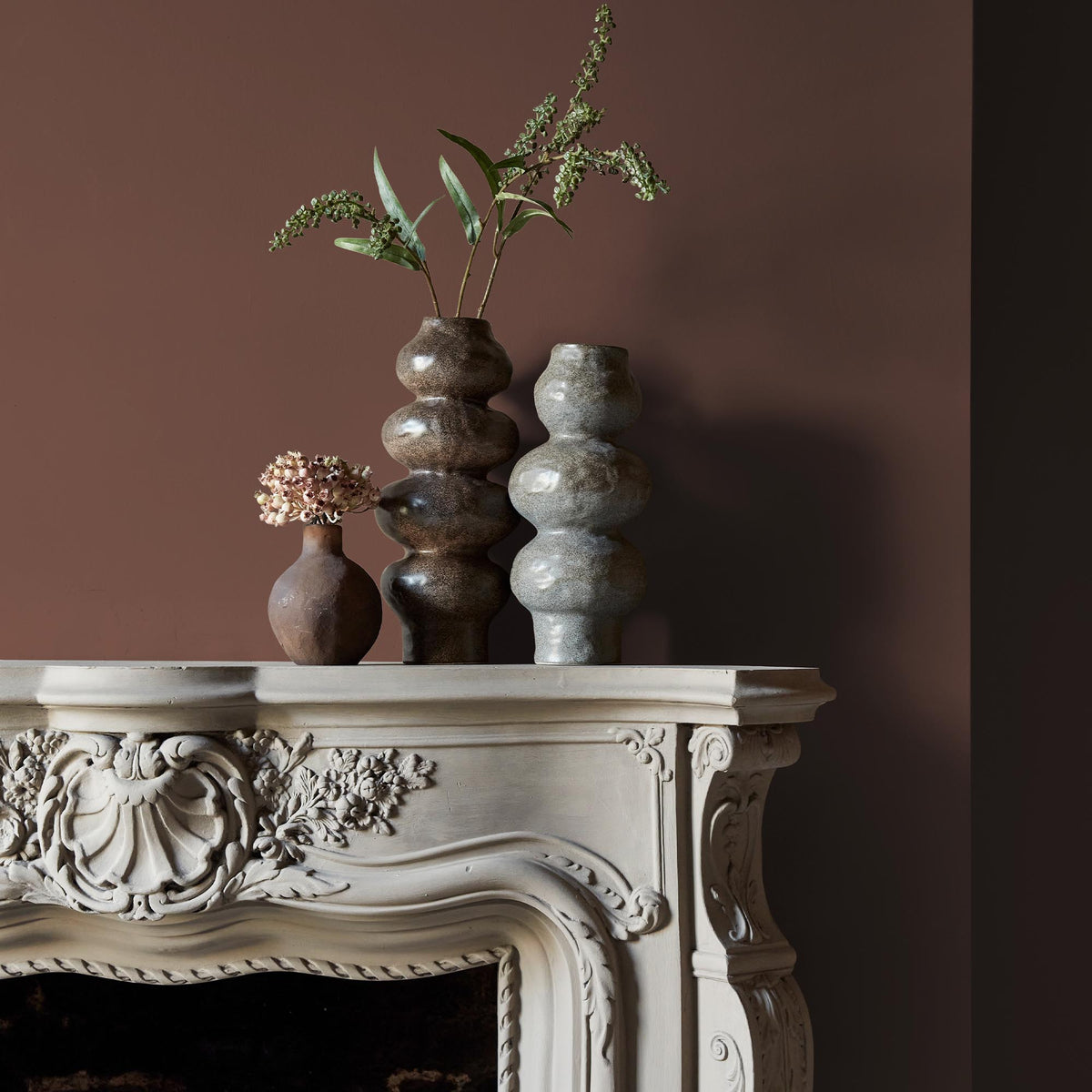 Three ceramic vases sitting on an intricate fireplace mantle in front of a wall in a rich, brown paint colour with a red undertone.