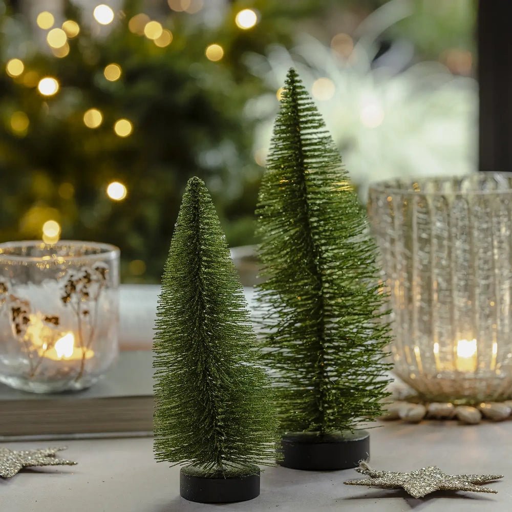 Orian green trees from the set of three stand on dark round bases, their tapered bottlebrush shapes framed by candleholders and warm festive lights.