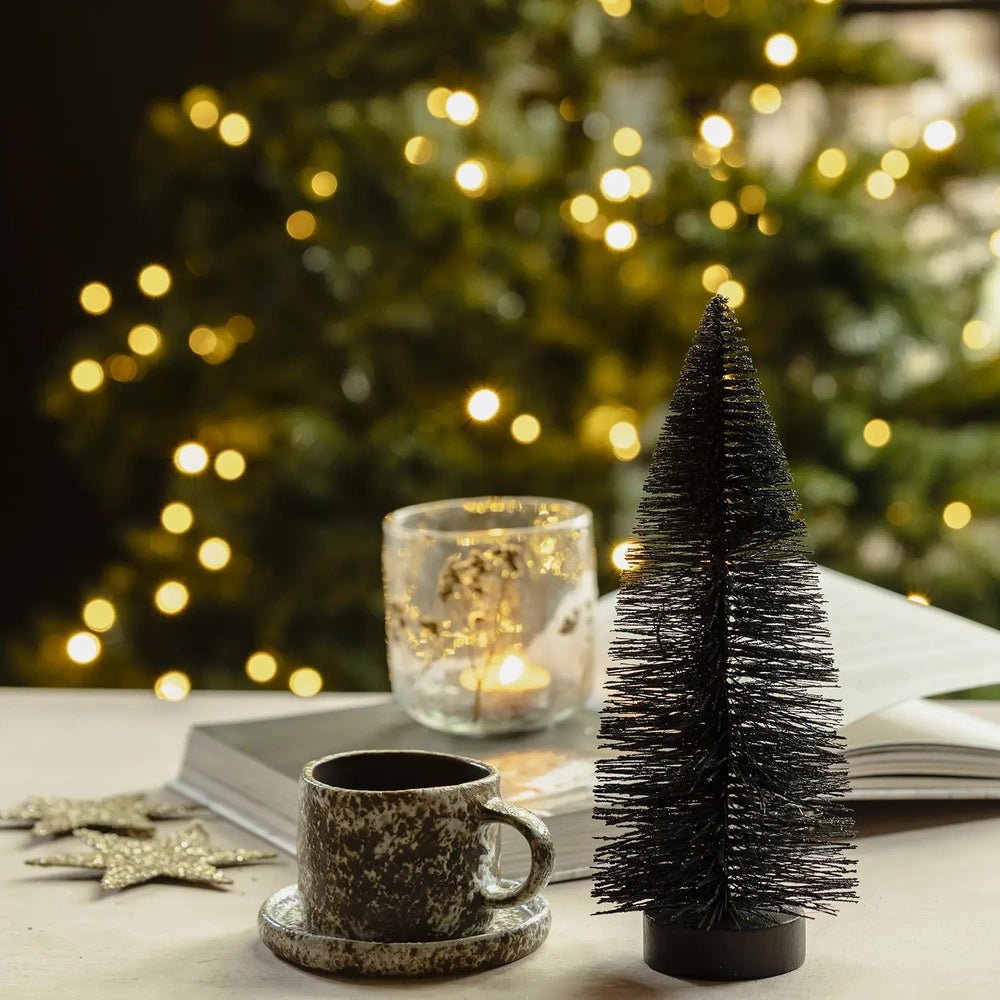 Orian medium midnight tree stands beside a mottled cup and saucer, its dark bottlebrush branches contrasting with a glowing candleholder and warm Christmas lights.