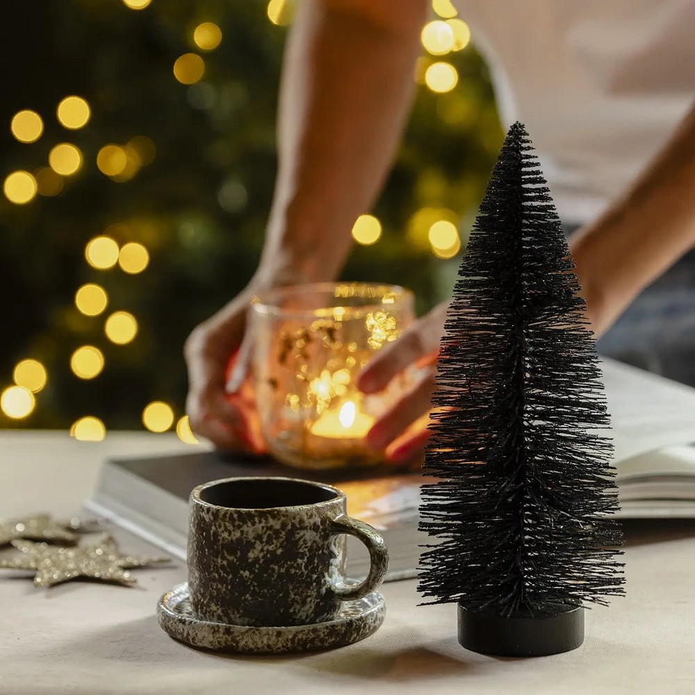 Orian medium midnight tree displays dark bottlebrush branches and a round base beside a cup and saucer, with hands arranging a glowing candleholder behind it.