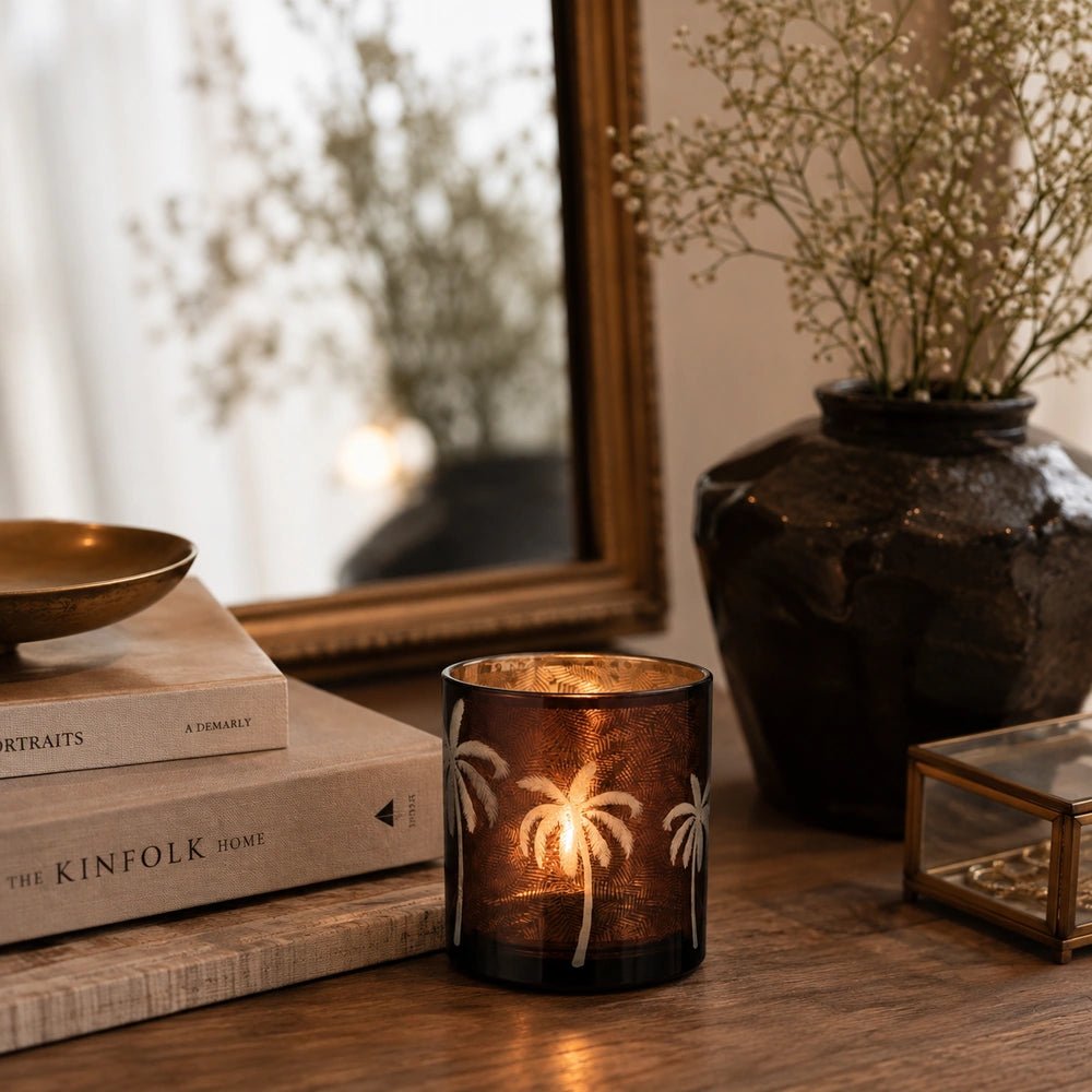 Palm Tree Glass Candle Holder glowing on a wooden console, showing etched palm motifs beside stacked neutral books, a dark vase and framed mirror.
