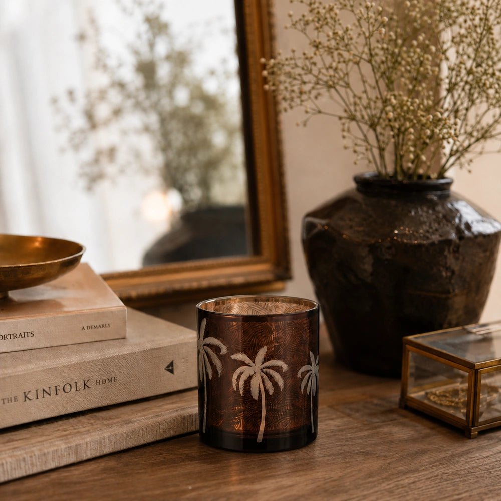 Palm Tree Glass Candle Holder in smoky amber glass, unlit on a wooden console beside stacked books, a dark vase, flowers and a framed mirror.