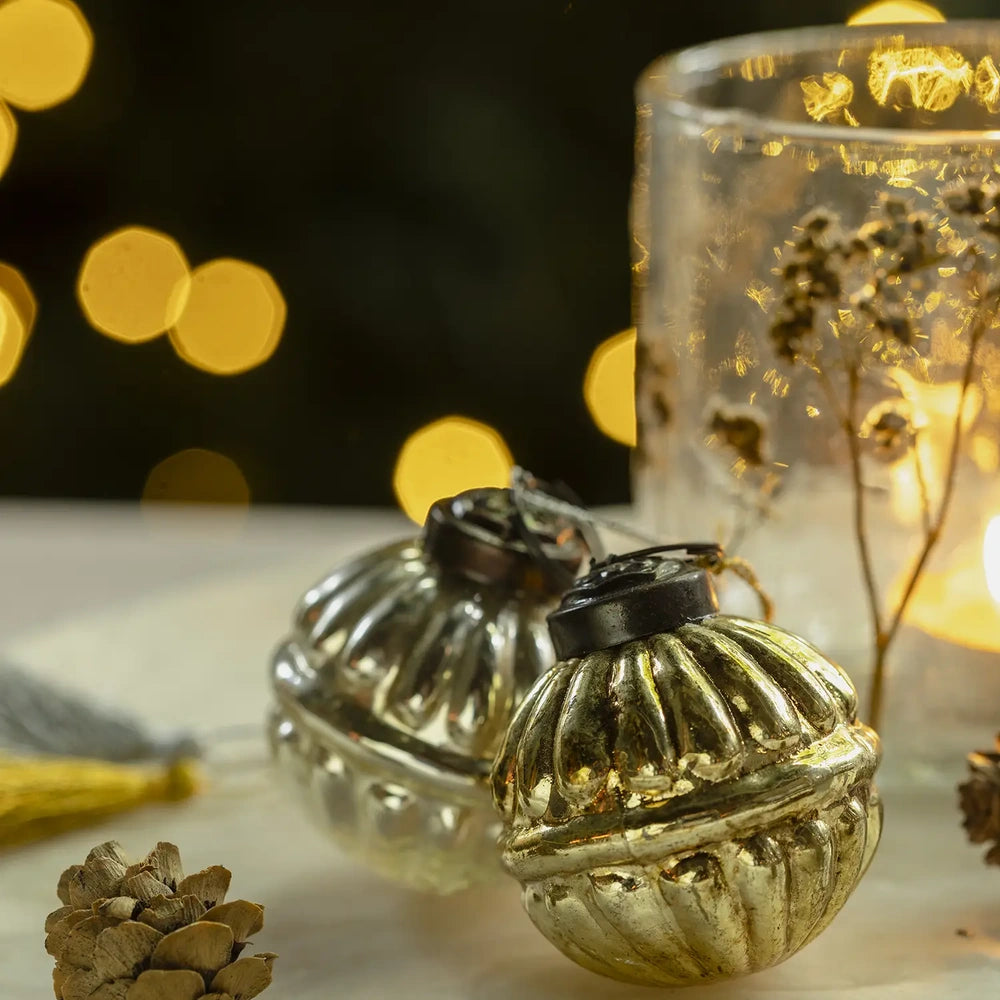 Pennington glass baubles have rounded ribbed shapes, reflective gold and silvery finishes, and dark hanging caps, resting beside a glowing candleholder on a festive tabletop.