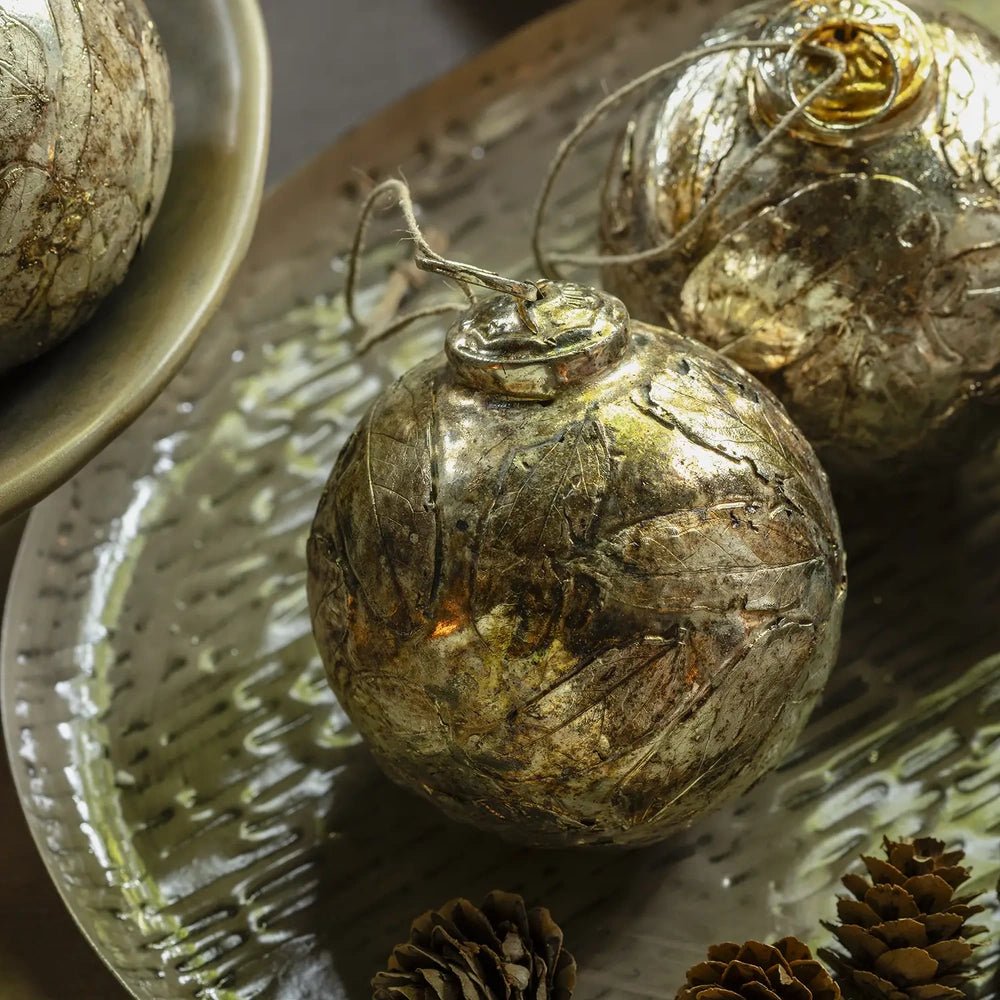 Perth antique silver leaf baubles have rounded shapes and mottled metallic surfaces, resting on a textured platter beside pine cones in a festive tabletop arrangement.