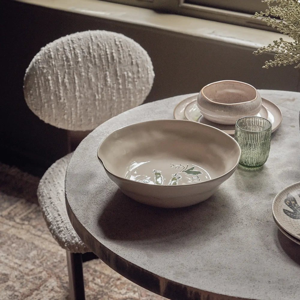 Quincy Ceramic Bowl sits on a round dining table beside a green glass and stacked dishes, with a cream glazed surface and softly uneven rim.