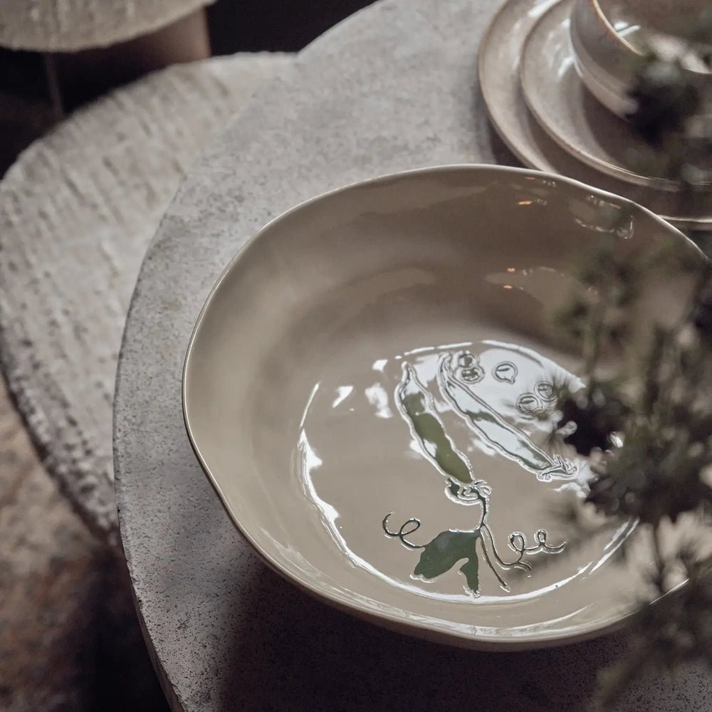 Quincy Ceramic Bowl displays green pea illustrations inside a shallow cream glazed form, viewed from above on a dining table beside glassware and soft textiles.