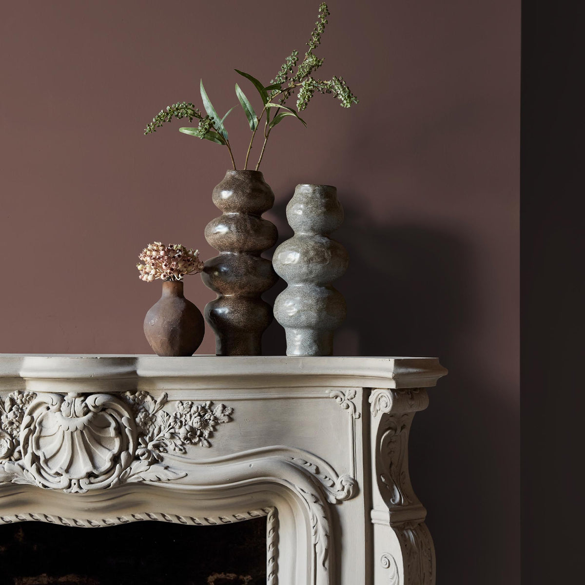 Three ceramic vases sitting on an intricate fireplace mantle in front of a wall in a warm, brown paint colour with a mauve undertone.