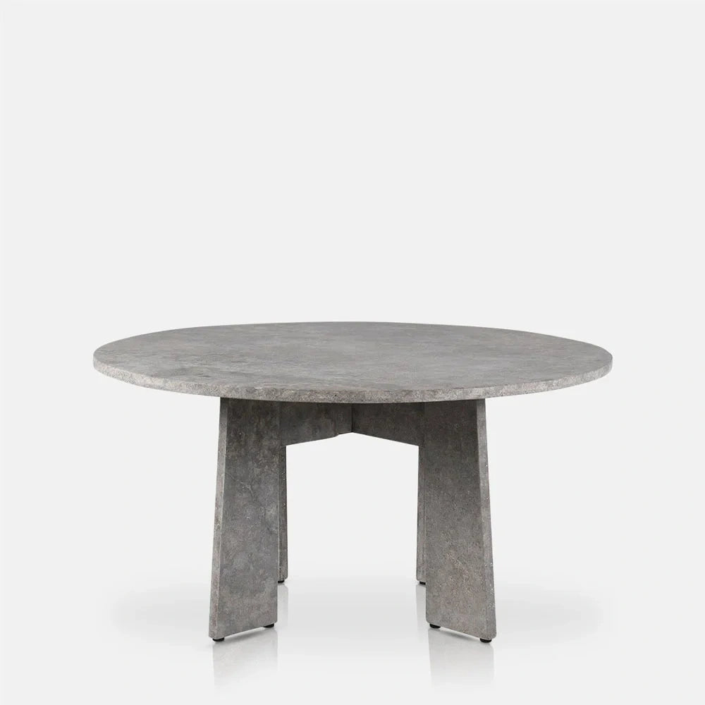 Randolph travertine dining table with a broad round grey top and angular slab legs, shown from the side to reveal its mottled stone surface.