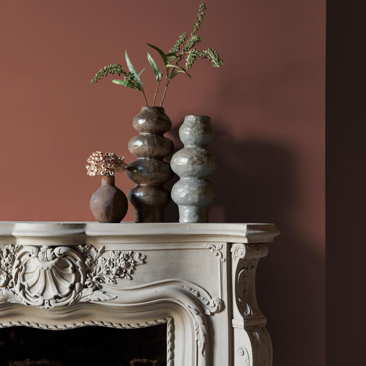 Three ceramic vases sitting on an intricate fireplace mantle in front of a wall in a bold, terracotta paint colour.