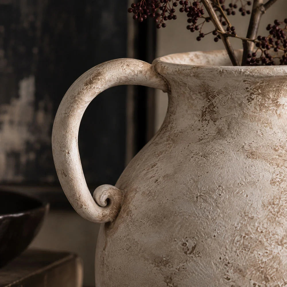 Romella ceramic vase shown close up, highlighting a curled handle, gently flared rim and weathered cream surface, with dark branches rising from the opening.