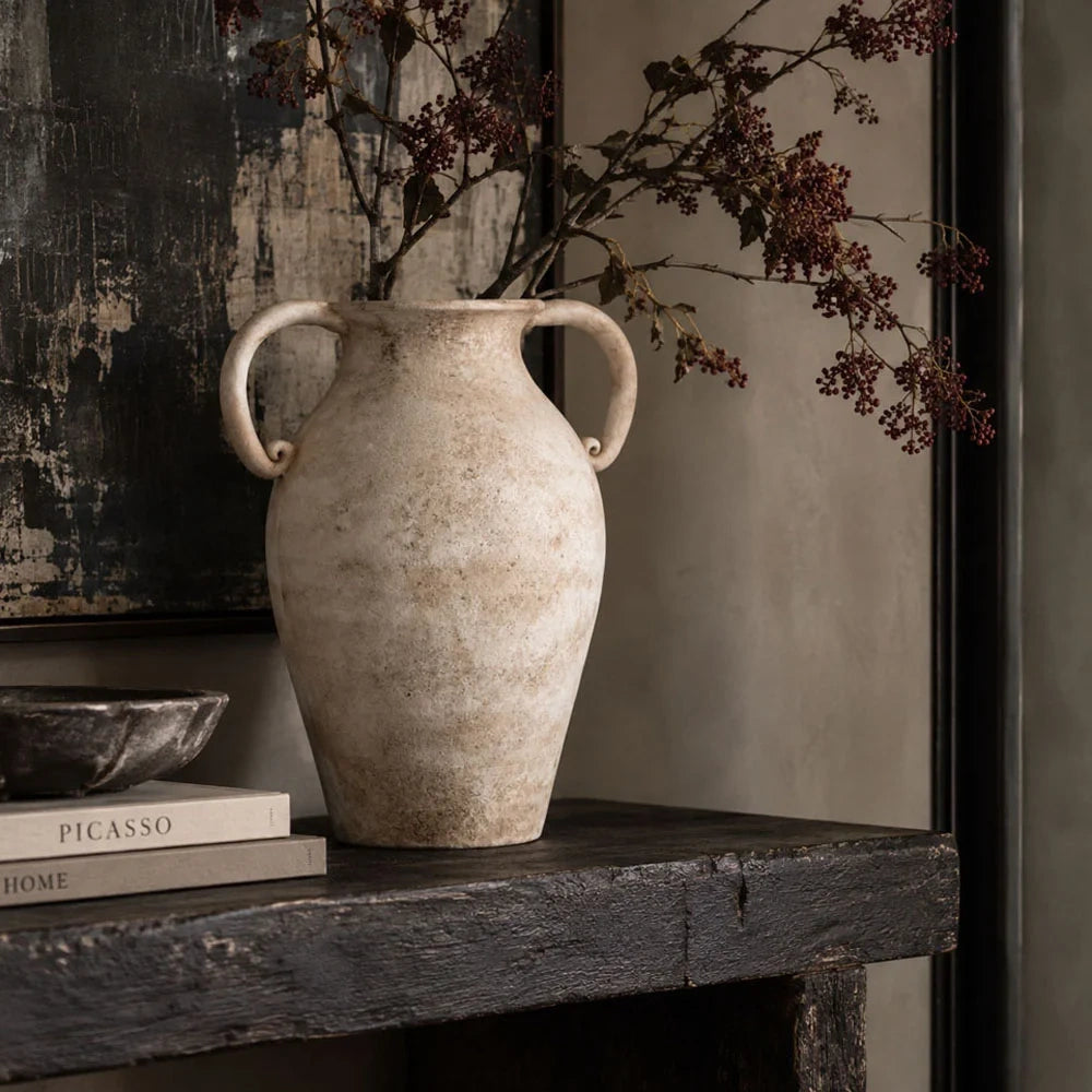Romella ceramic vase with a tall rounded body, two curled handles and a mottled cream surface, holding branches on a dark console beside stacked books.