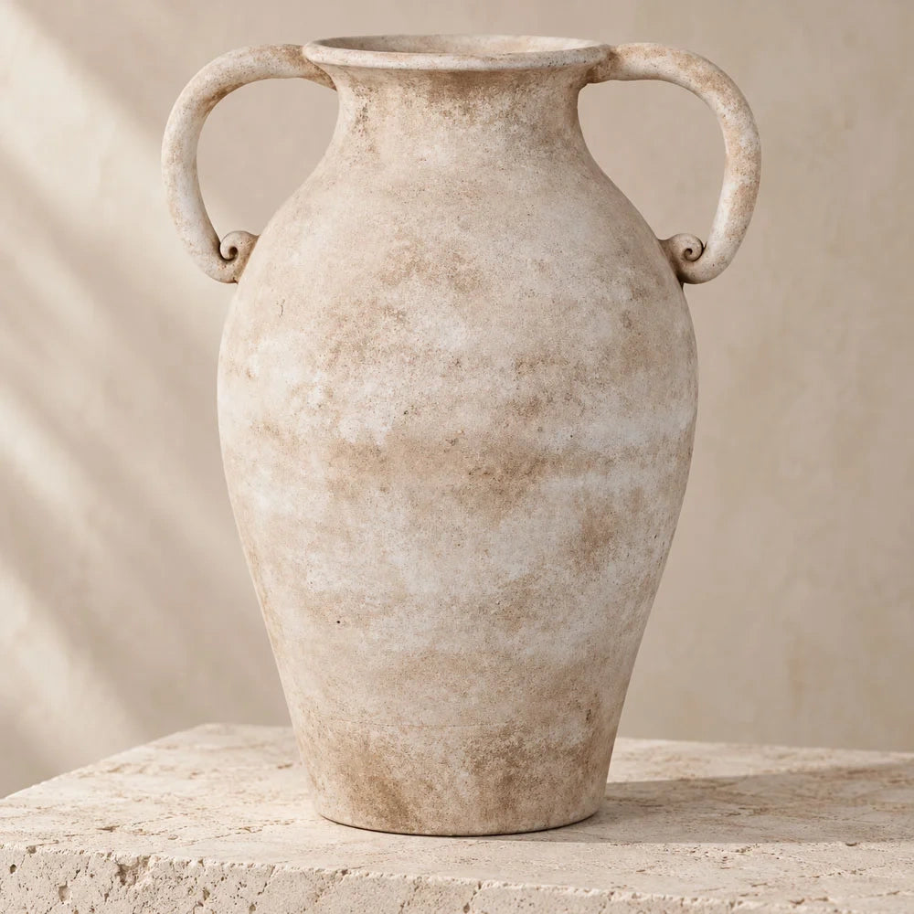 Romella ceramic vase with two curved handles, a tapered base and a softly mottled cream finish, displayed on a pale stone plinth against a neutral wall.
