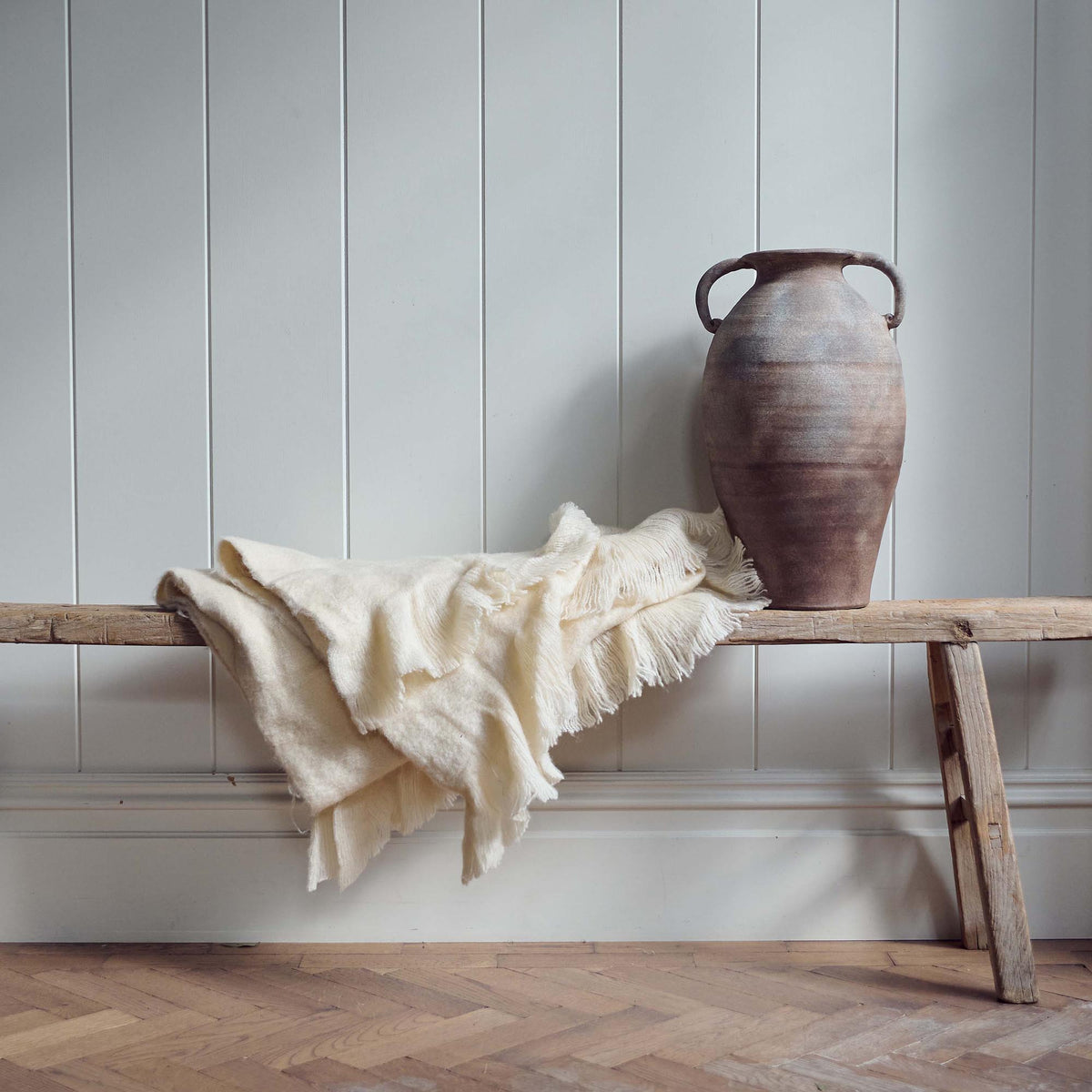 The Borough Beige Throw, a soft cream coloured throw with fringed ends on a low wooden bench next to a terrracotta vase.