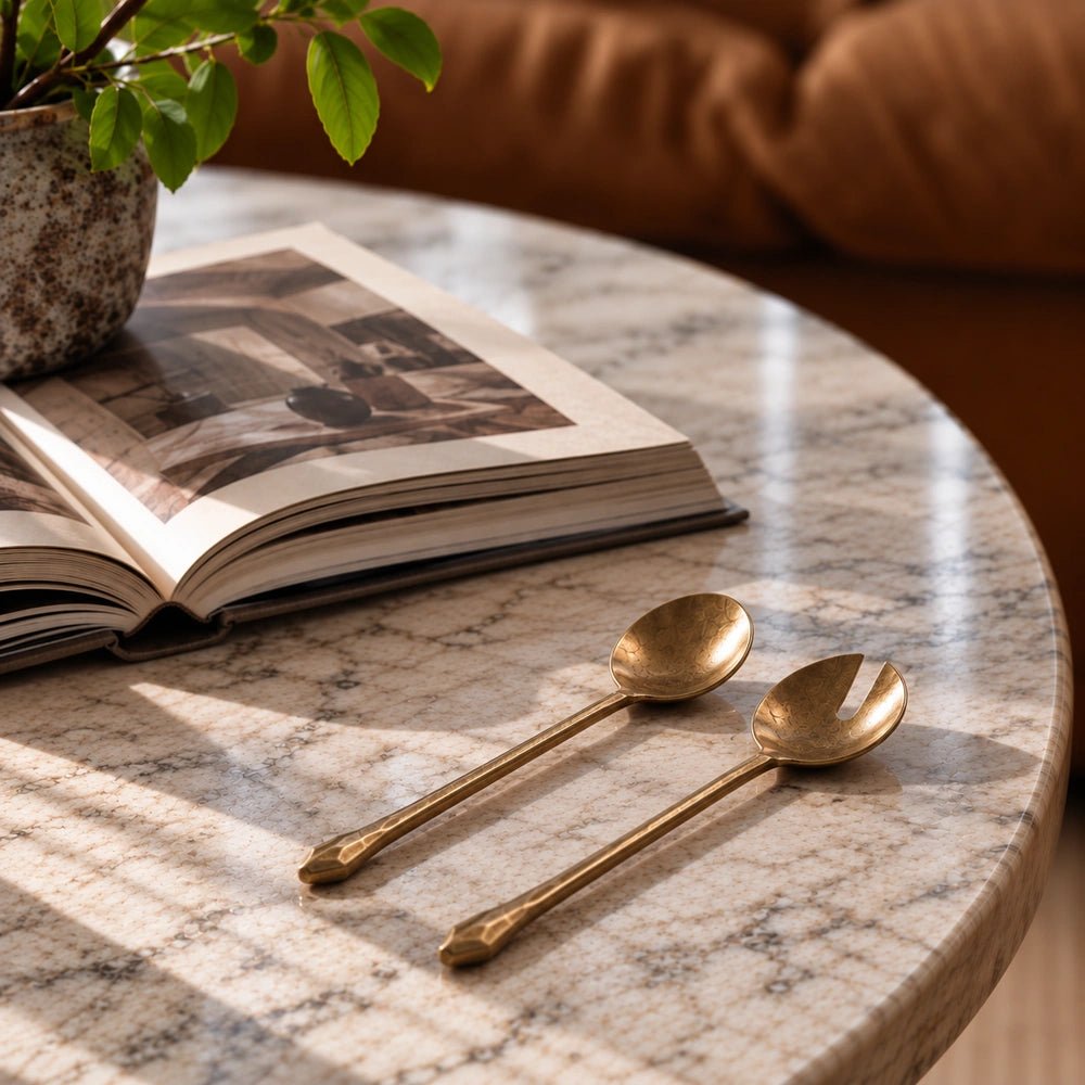 Rooke Metal Salad Servers in warm brass tones, laid on a marble table beside an open book and leafy plant in soft natural light.