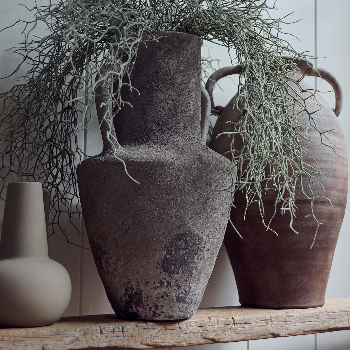 The Rowan Terracotta Vase, an angular grey amphora shaped vase with two handles holding some wispy faux trailing green stems.