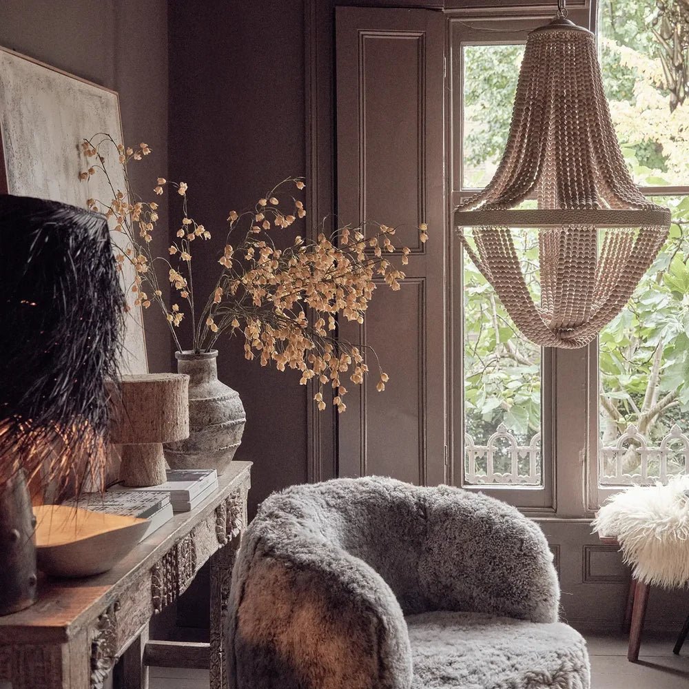 Roxham beaded chandelier with cascading bead strands and a circular central band, hanging above a textured armchair beside a window and a console with branches.
