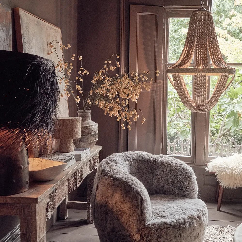 Roxham beaded chandelier with draped strands and a wide central band, suspended beside a tall window above a textured armchair in a warm living room.