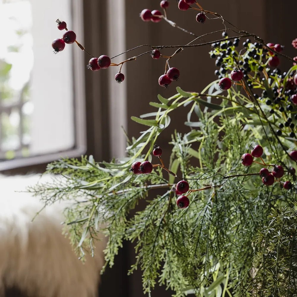 Ruston artificial bouquet shows slender red berry branches above fine green foliage, with trailing stems creating an airy arrangement beside a softly lit window.