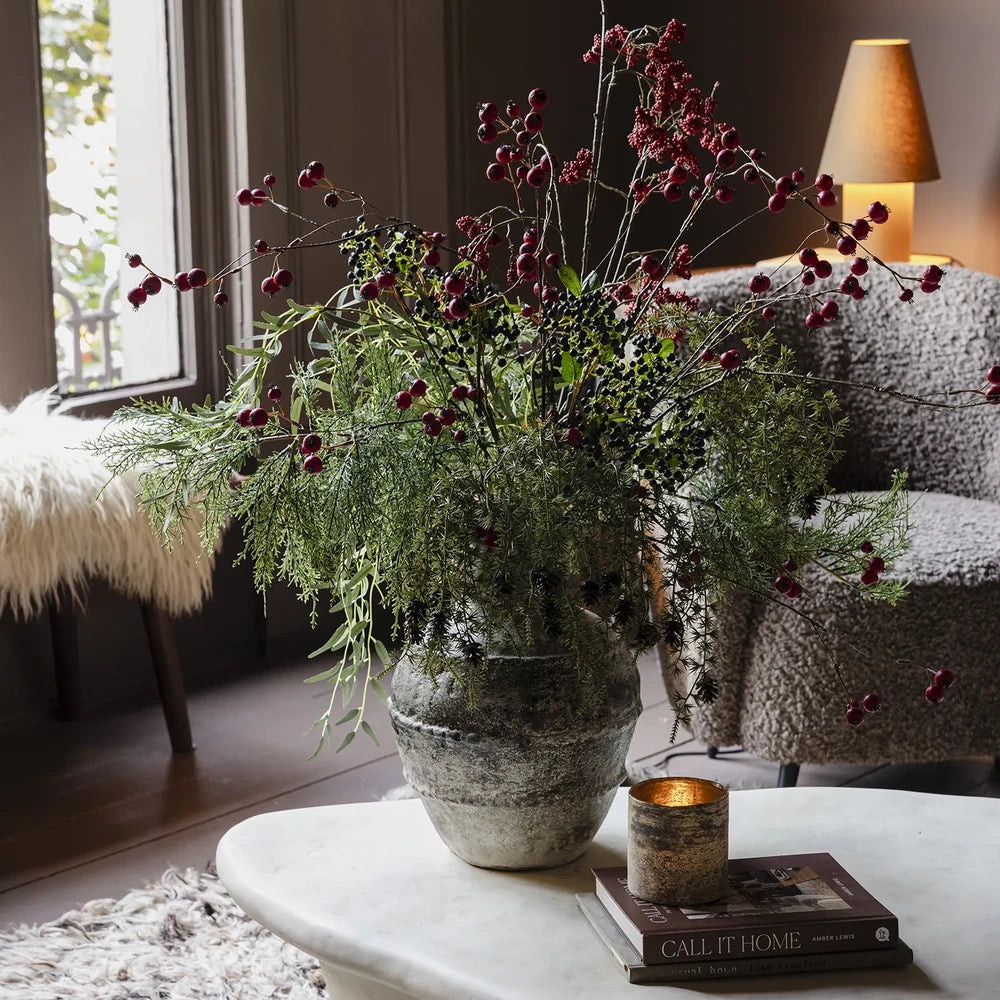 Ruston artificial bouquet combines airy green foliage and tall red berry branches in a textured vase, displayed on a coffee table beside a sofa.
