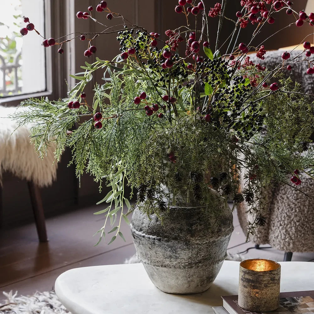 Ruston artificial bouquet spreads green foliage and red berry branches from a textured rounded vase, styled beside a candle on a living room coffee table.