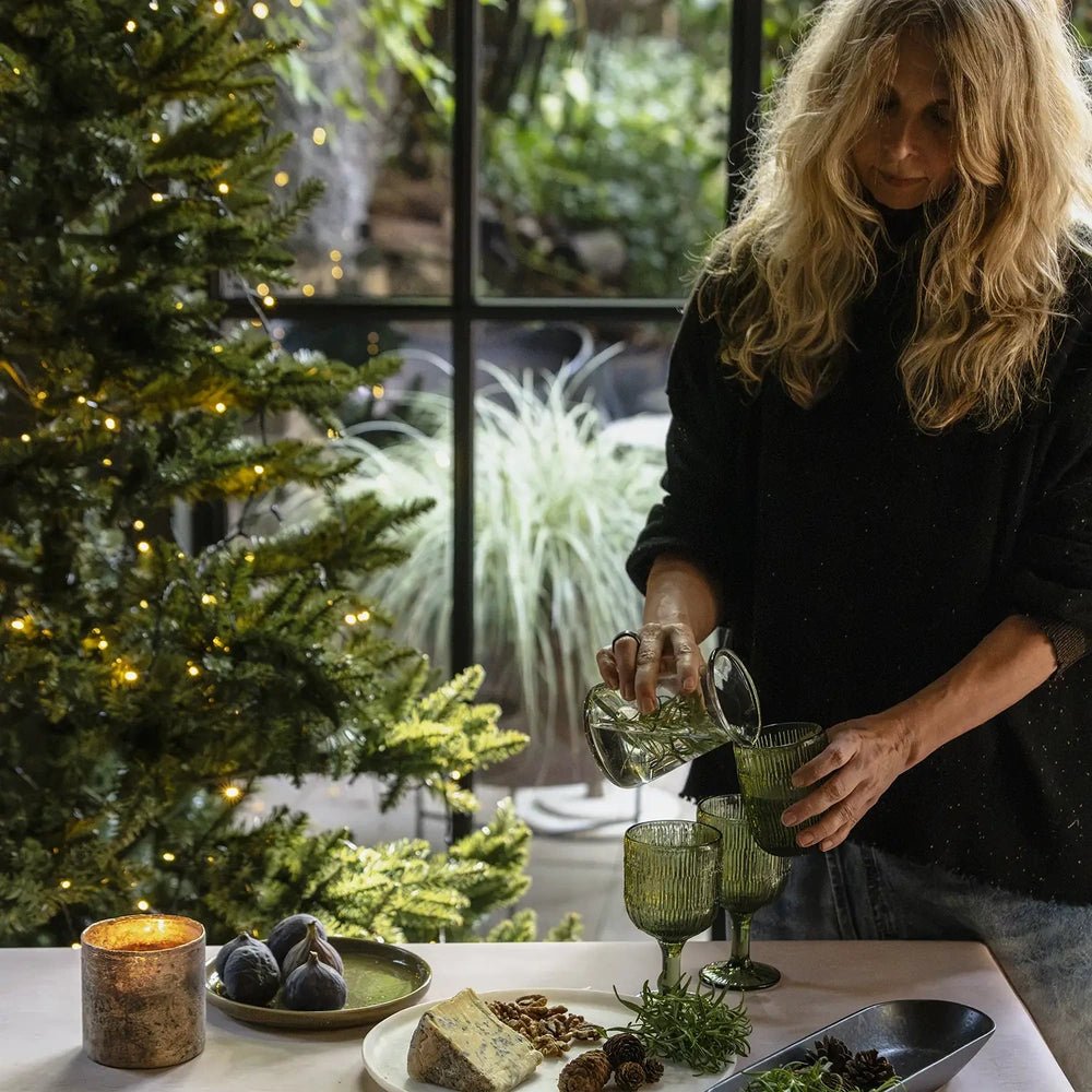 Salome round platter displays cheese, walnuts and greenery beside textured green glasses, with a person pouring water at a table near a glowing Christmas tree.