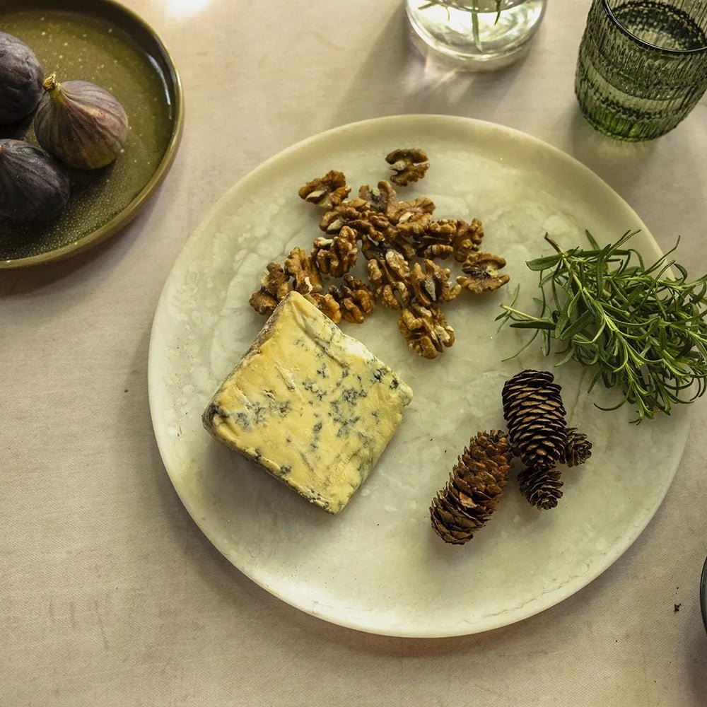 Salome round platter has a pale mottled finish and gently raised rim, holding blue cheese, walnuts, pine cones and greenery in an overhead table setting.