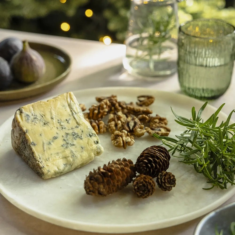 Salome round platter shows its pale textured surface and raised rim beneath blue cheese, walnuts and pine cones, with green glassware in the softly blurred background.