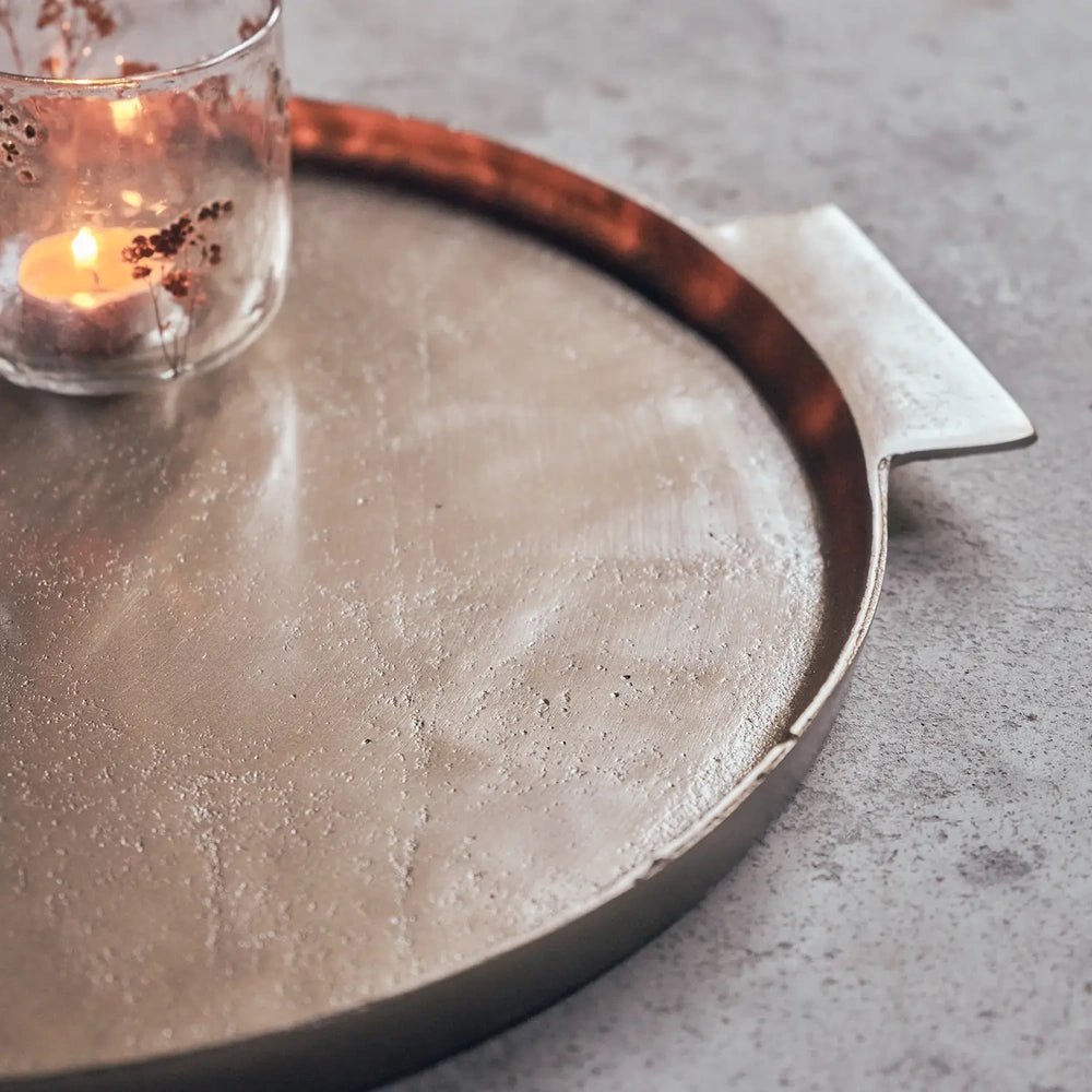 Sandford metal decorative tray shown close up beside a candle, highlighting its reflective warm toned surface, shallow circular rim and broad flat rectangular handle.