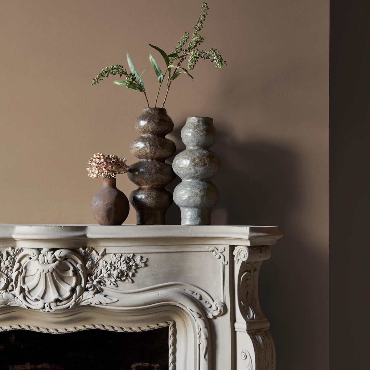 Three ceramic vases sitting on an intricate fireplace mantle in front of a wall in a soft, brown paint colour with golden undertones.