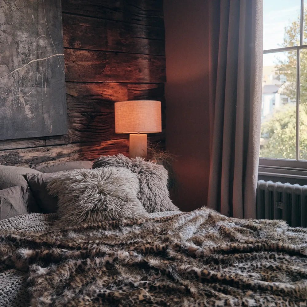 Savannah throw with a plush animal patterned surface in brown and cream, spread across a sofa beneath textured cushions beside a brick wall and table lamp.
