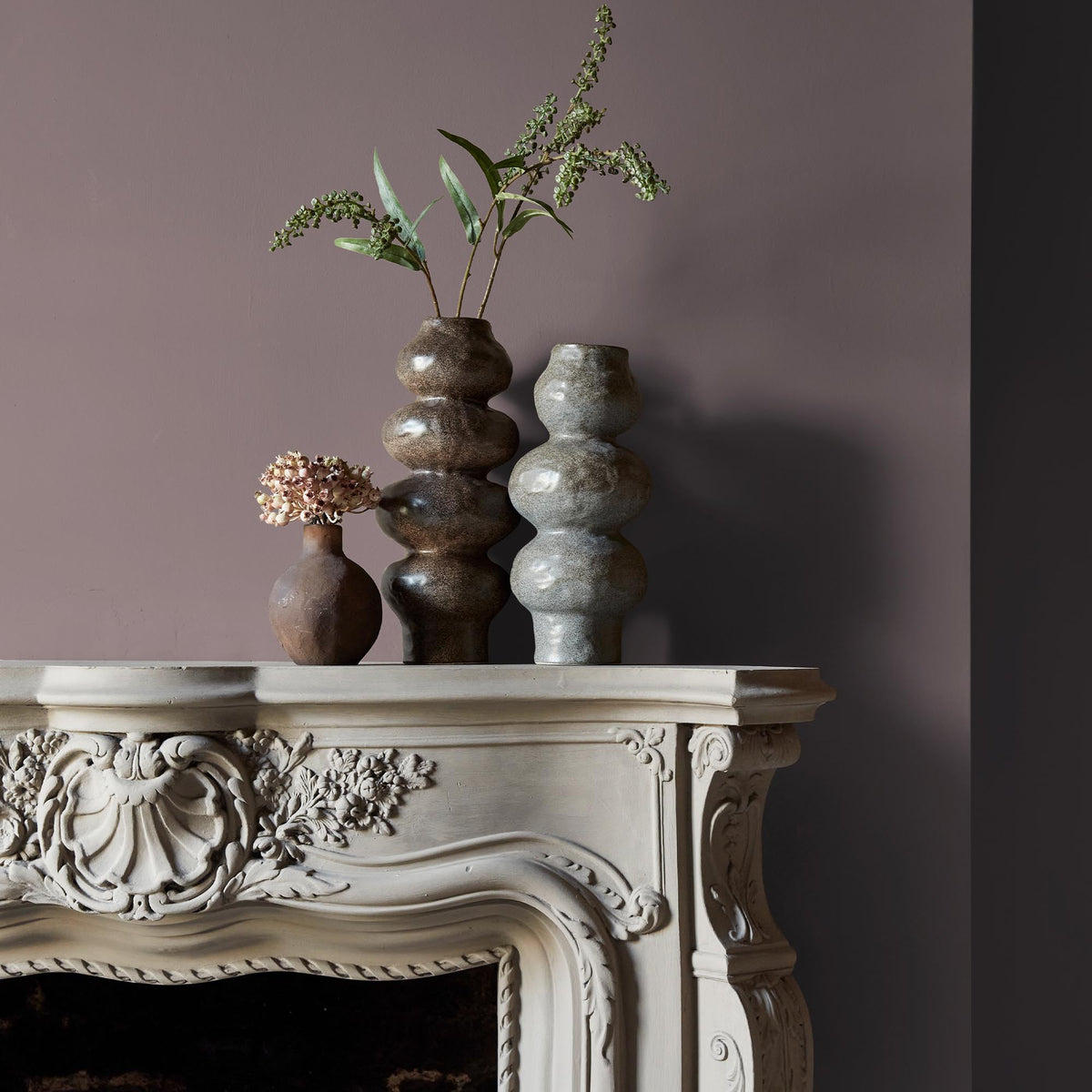 Three ceramic vases sitting on an intricate fireplace mantle in front of a wall in a warm, dusky purple paint colour.
