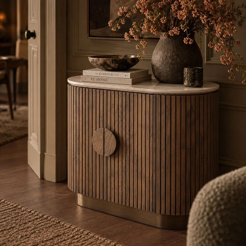 Sheridan wooden cabinet with curved fluted doors, a round handle and pale top, viewed at an angle beside textured seating with branches and books above.