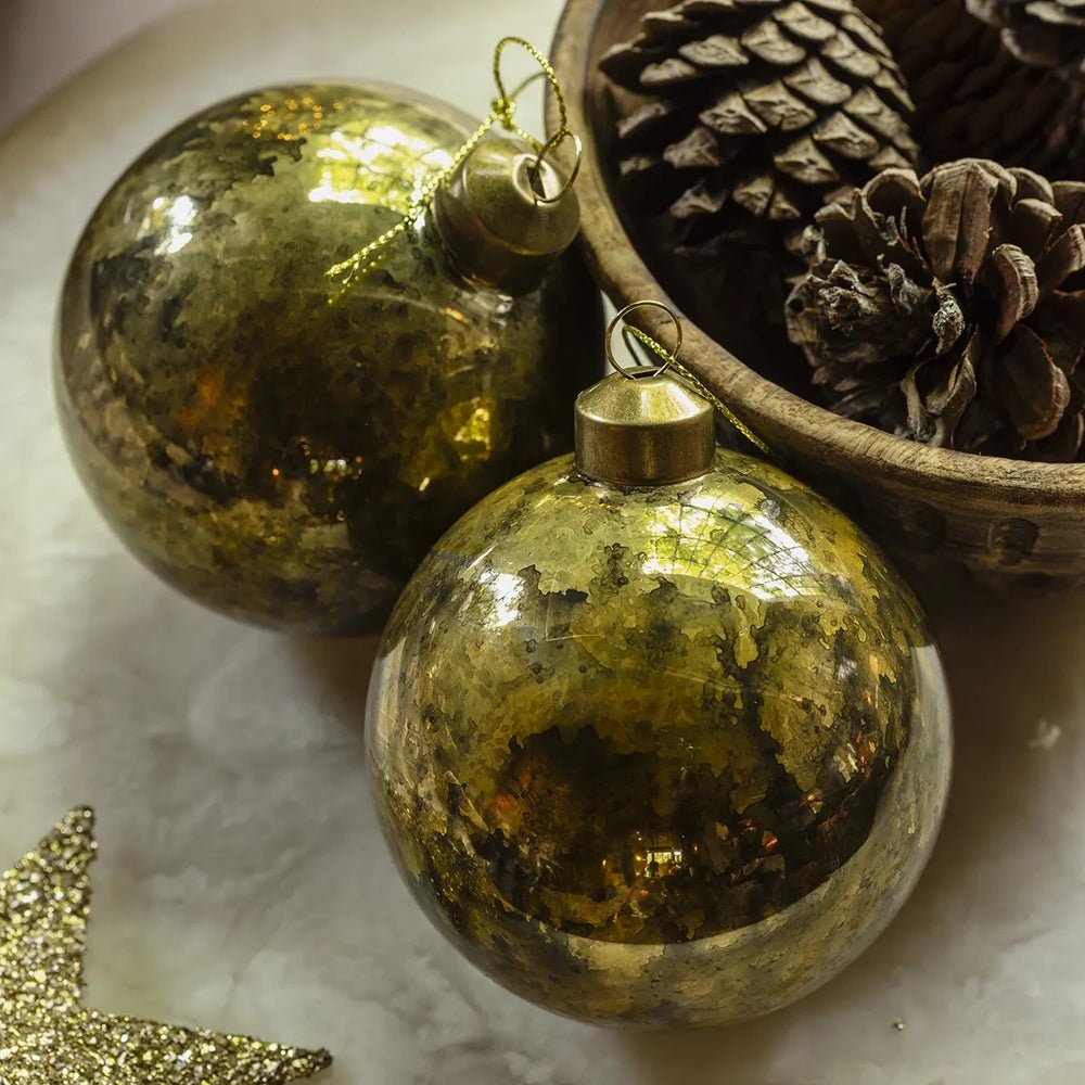 Silas antique gold baubles show contrasting mottled patches across rounded reflective surfaces, arranged as a pair beside a bowl of pine cones on a pale tabletop.