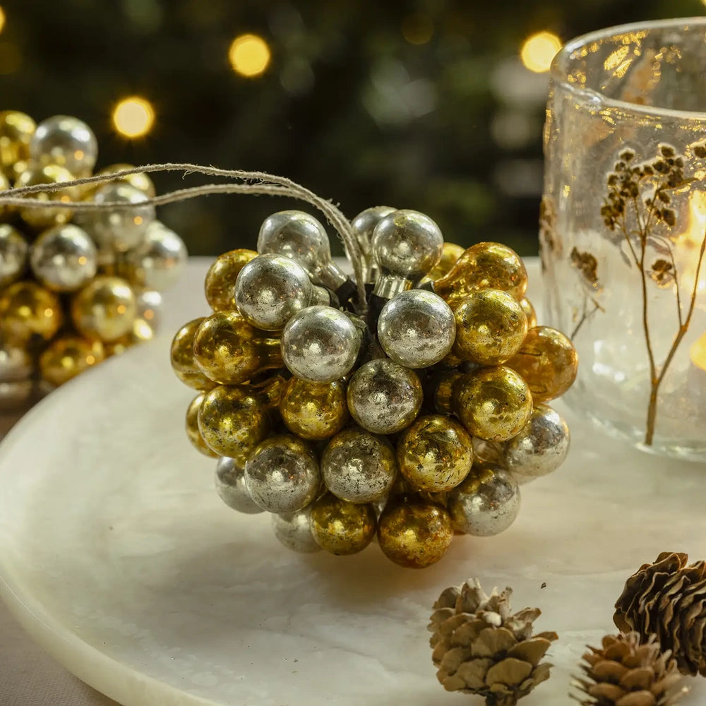 Silvero glass bauble forms a rounded cluster of small gold and silver spheres, resting on a pale platter beside a glowing decorative candleholder.