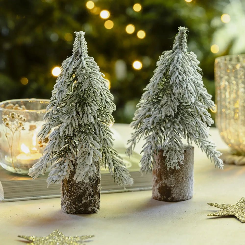 Snowy tree decorations display layered green branches covered with a white snowy finish, standing on rustic brown bases beside candleholders in a festive tabletop setting.