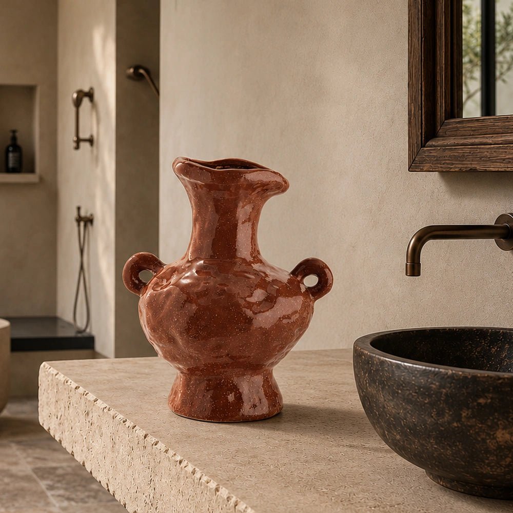 Sylvan Stoneware Vase with sculptural handles and glossy brown finish, displayed on a pale stone bathroom console beside a dark basin in warm natural light.