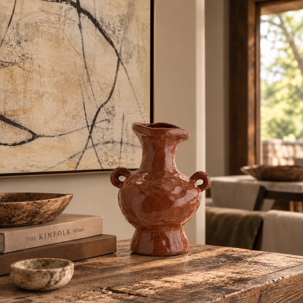 Sylvan Stoneware Vase with sculptural handles and glossy brown finish, styled on a textured coffee table beside stacked books and decorative bowls in warm light.