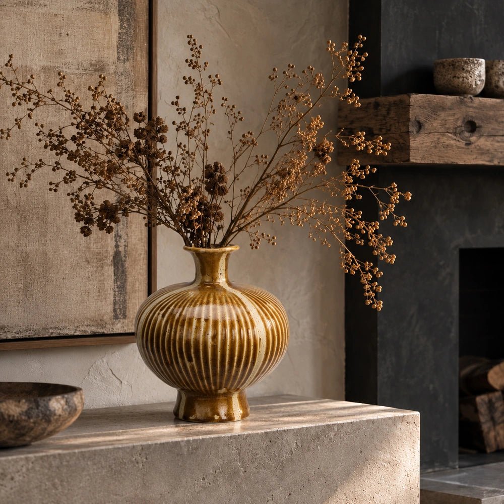 Tallis Stoneware Vase with ribbed ochre surface, filled with dried branching stems on a pale stone console beside textured artwork and a dark fireplace.