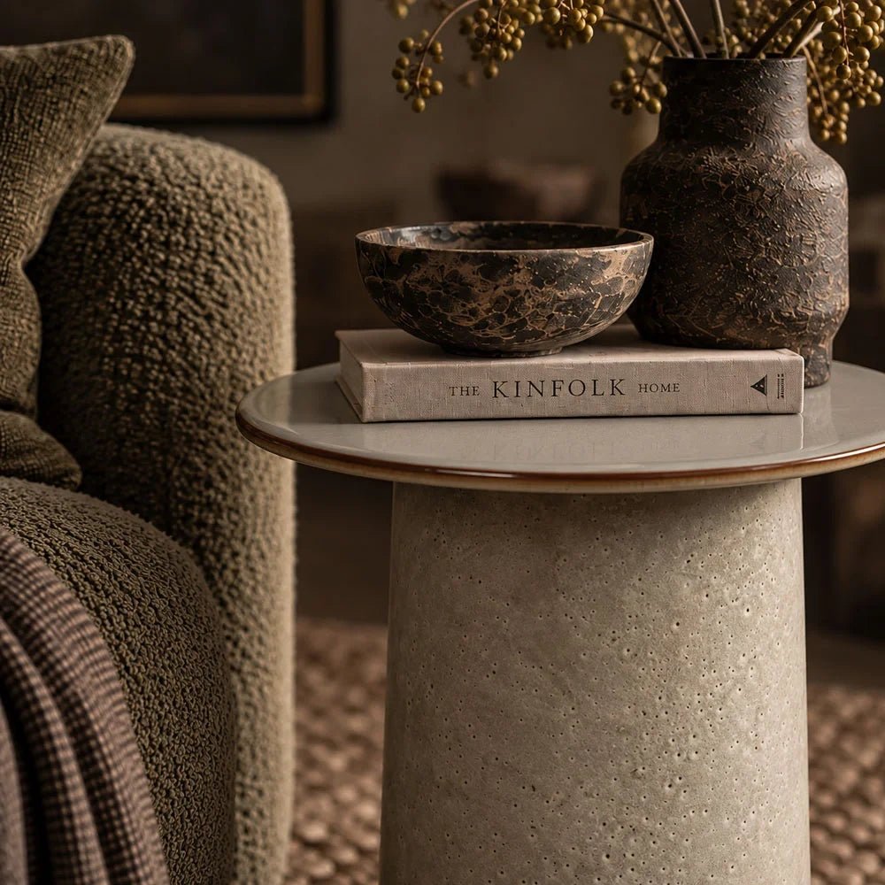 Thacker stoneware side table with a round pale top and textured cylindrical base, holding books and decorative vessels beside a green textured armchair.