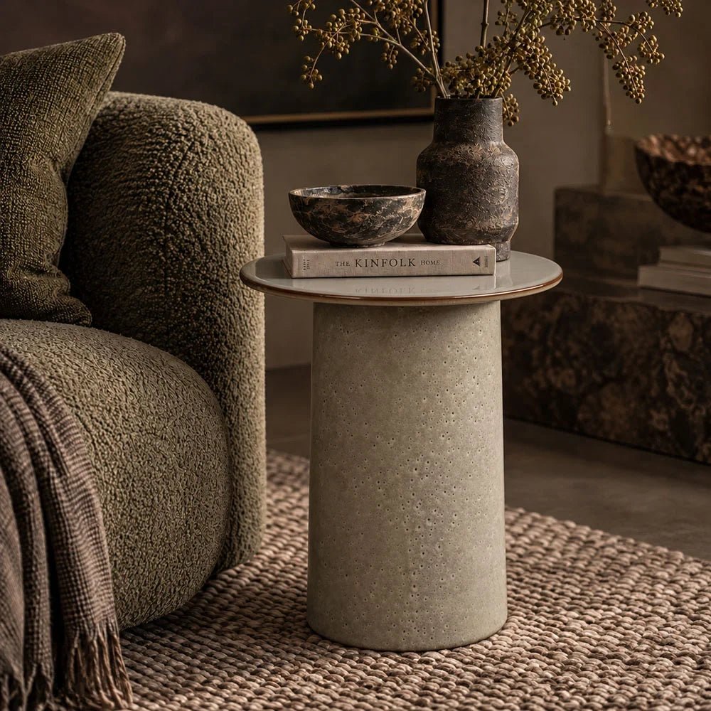 Thacker stoneware side table with a pale round top and lightly mottled cylindrical base, styled beside an armchair with books and a vase on a woven rug.