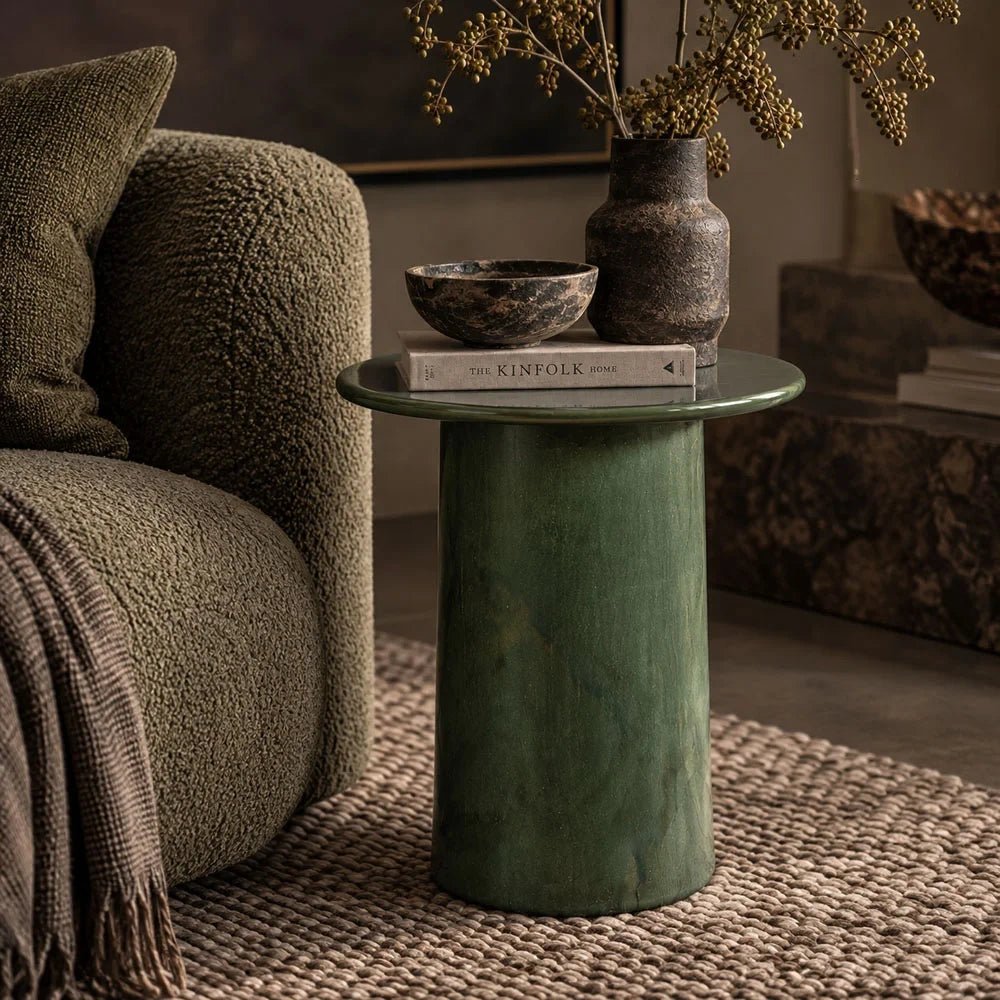 Thayer stoneware side table with a glossy green round top and cylindrical base, styled with books and decorative vessels beside a textured armchair on a woven rug.