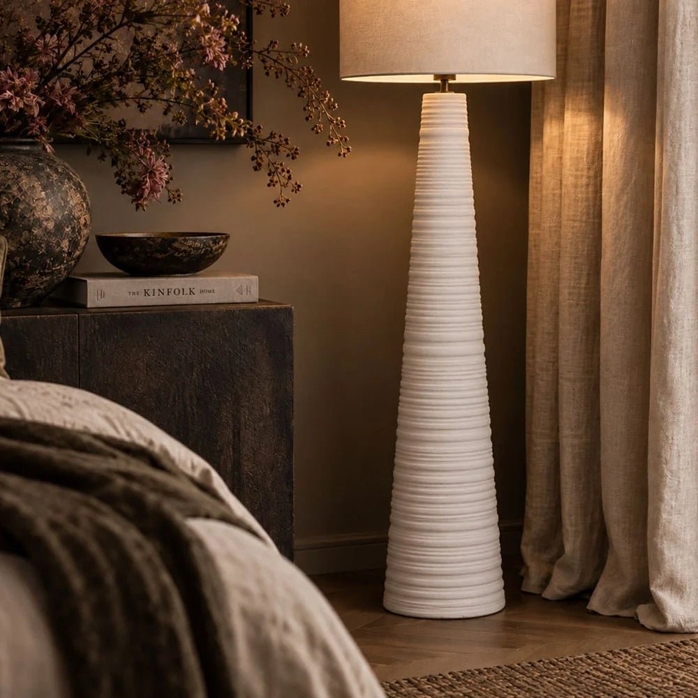 Vanderbilt ceramic floor lamp with a tall white ribbed base and pale fabric shade, glowing beside a bed, neutral curtains and a console with branches.