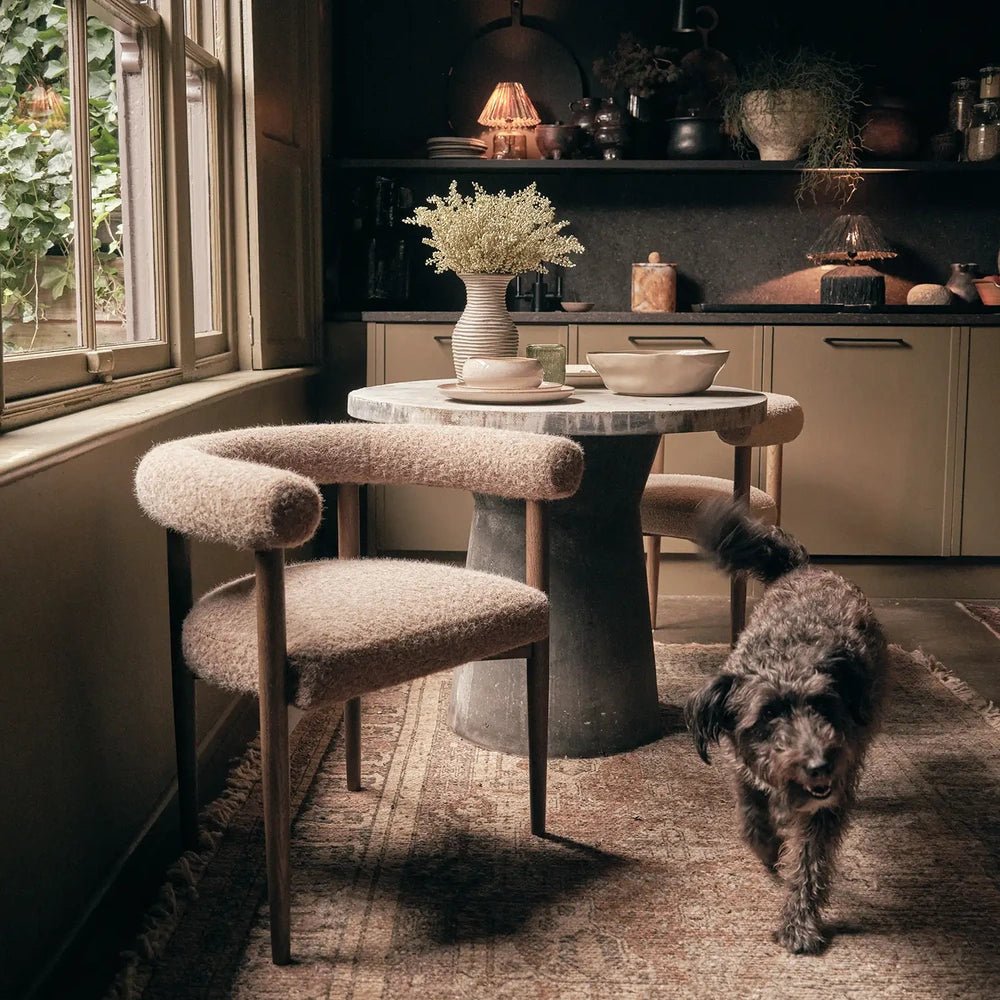 Virgil upholstered dining chairs with curved padded arms and taupe textured fabric surround a round table, with a dark dog standing in the dining room.
