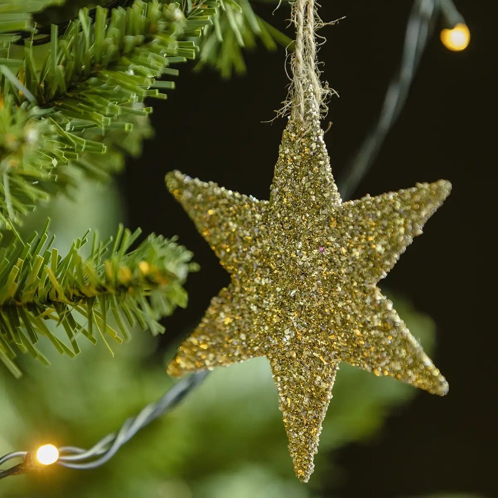 Volta glitter star bauble displays dense gold glitter across a flat five-pointed surface, suspended beside green Christmas tree branches with a small warm light nearby.