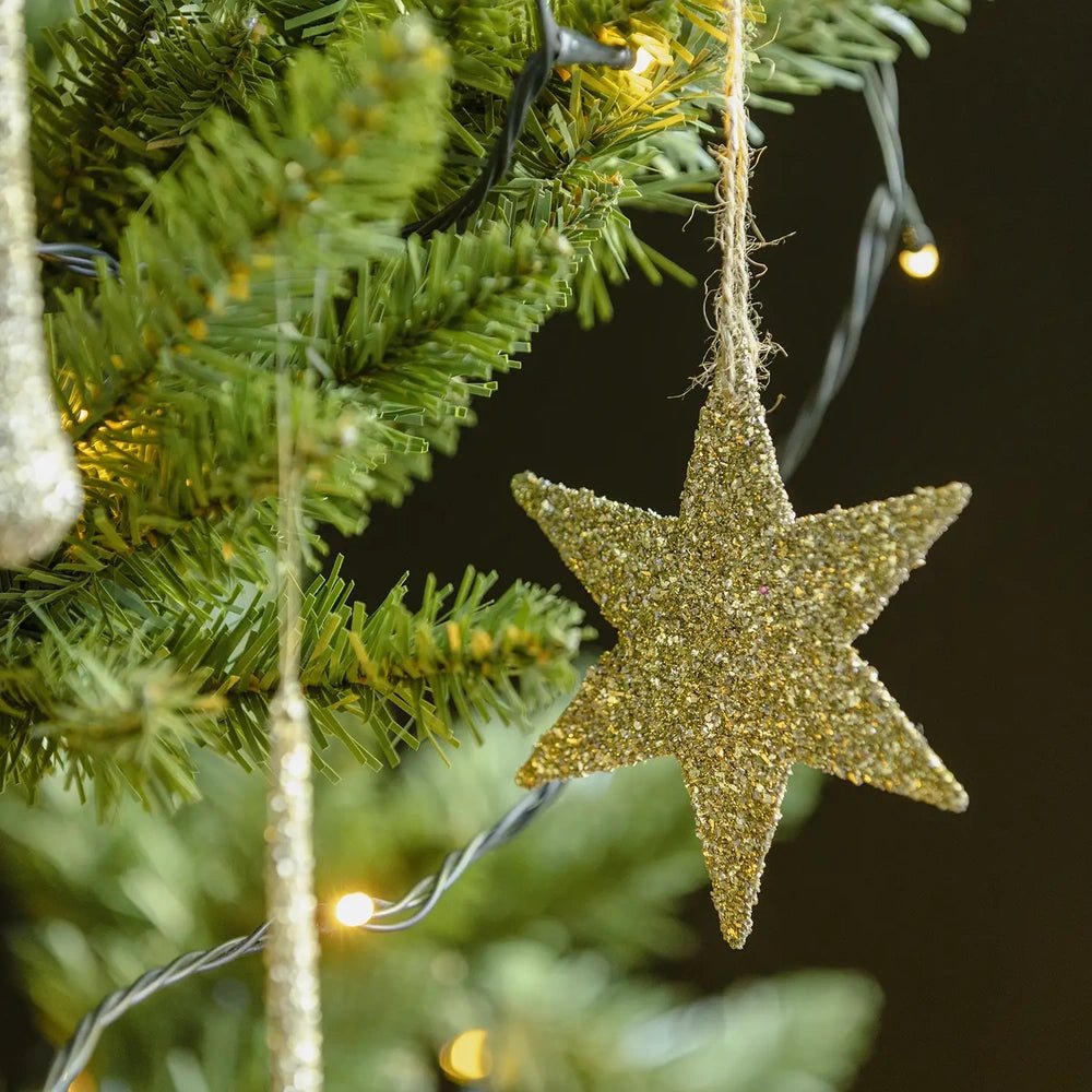 Volta glitter star bauble shows a flat five-pointed shape covered in gold glitter, hanging from a cord beside green tree branches and warm lights.