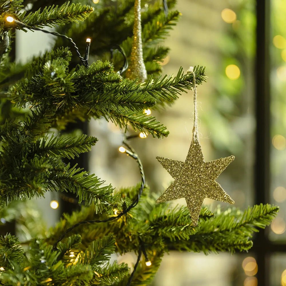 Volta glitter star bauble hangs from a gold cord among green Christmas tree branches, its flat five-pointed shape framed by softly glowing warm lights.