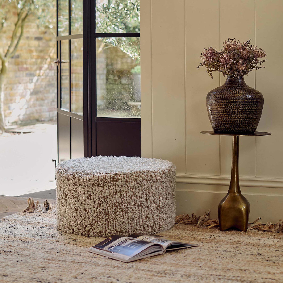 The Walker Boucle Pouffe, a circular pouffe in warm brown with cream boucle accents in a bright sunny lounge room.